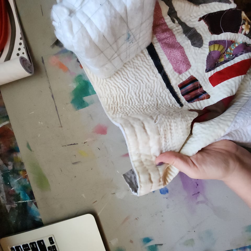 Top-down view of Sarah's hand as she puts the final stitches in Meet Me at the Corner, a patchwork quilt consisting of many scraps of fabric sewn onto an off-white background. Parts of Sarah's desk are visible, including a paperback book and one corner of her laptop.