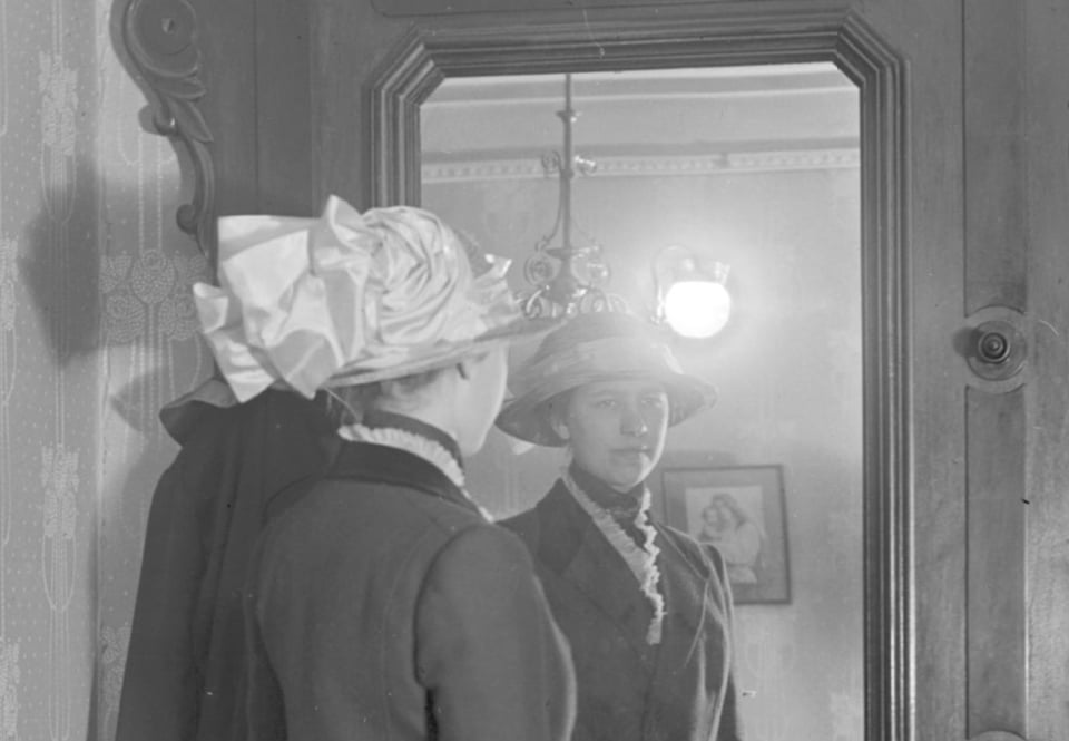 An old black-and-white photo of a well-dressed woman with an elaborate satin hat looking in the mirror, with a picture of mother and child on the wall in the background.