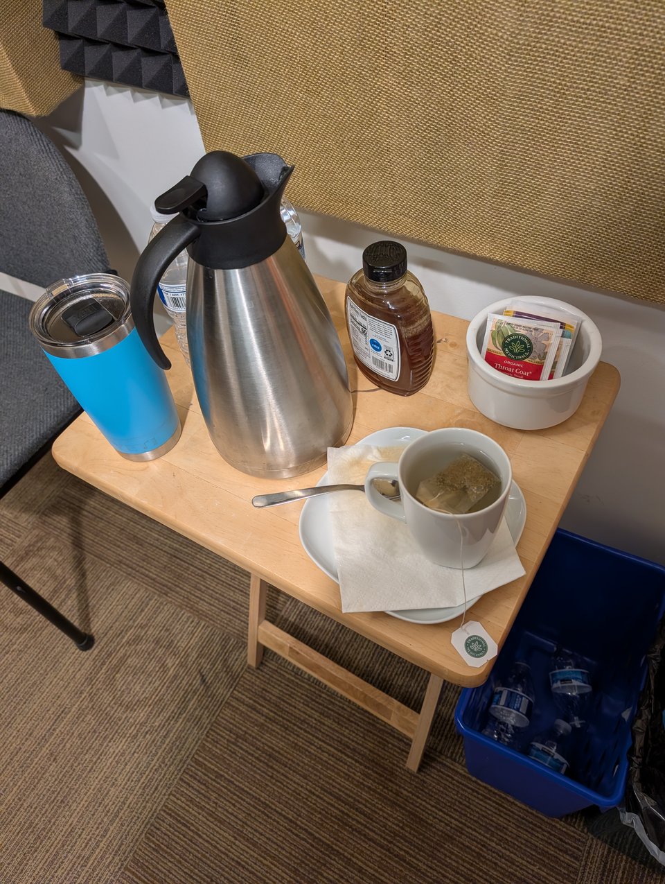 A photograph of a small table with bottled water, a travel coffee mug, a carafe of hot water, a bunch of decaffeinated tea, and honey.
