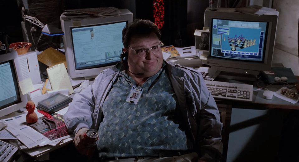 A screenshot of the actor Wayne Wright in the movie Jurassic Park, sitting facing the camera. Behind him is a desk with three large 1990s computer monitors; the desk is cluttered with paper and keyboards and a small folded up digital assistant