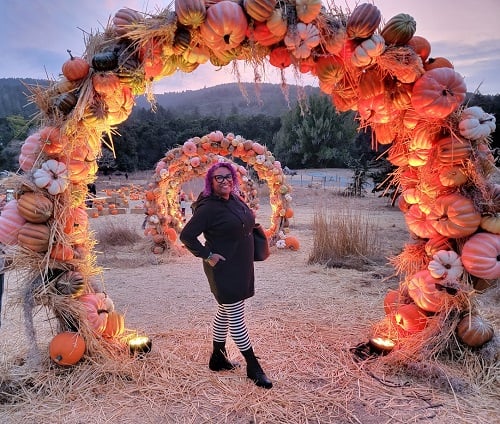 Patricia in a black dress and striped tights standing under an archway of pumpkins