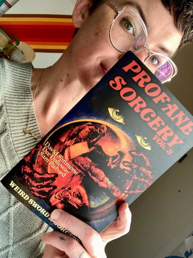 D, a white woman in a green sweater and gold-frame glasses, holding a copy of Profane Sorcery partially in front of her face.
