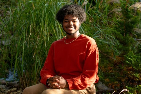 A young man with curly hair sits outdoors, wearing a red sweatshirt and smiling warmly. He is positioned near tall green plants and rocks, suggesting a natural, serene environment.