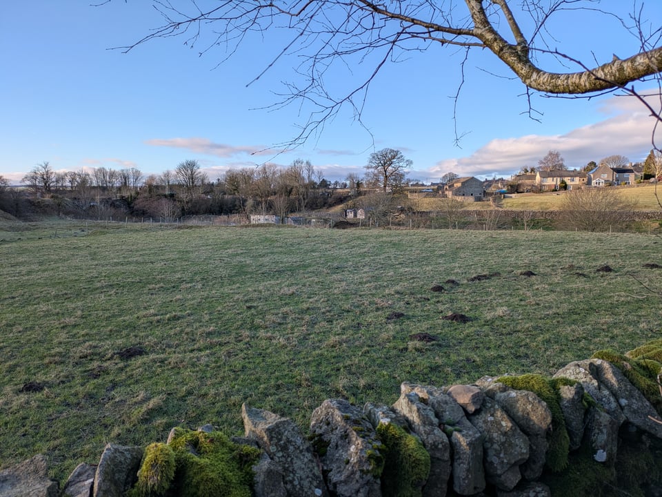 A shaded field under a blue sky, beyond a mossy dry stone wall. The sun touches some houses in the distance.
