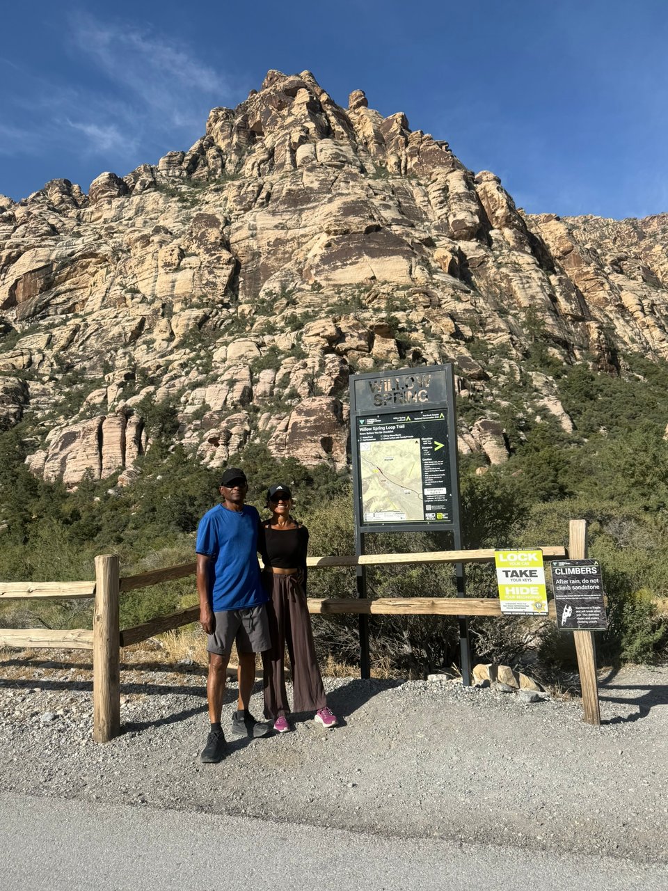 Photo of my dad and I standing in front of the Willow Spring trailhead sign with towering red and cream sandstone cliffs rising behind them beneath a clear blue sky.