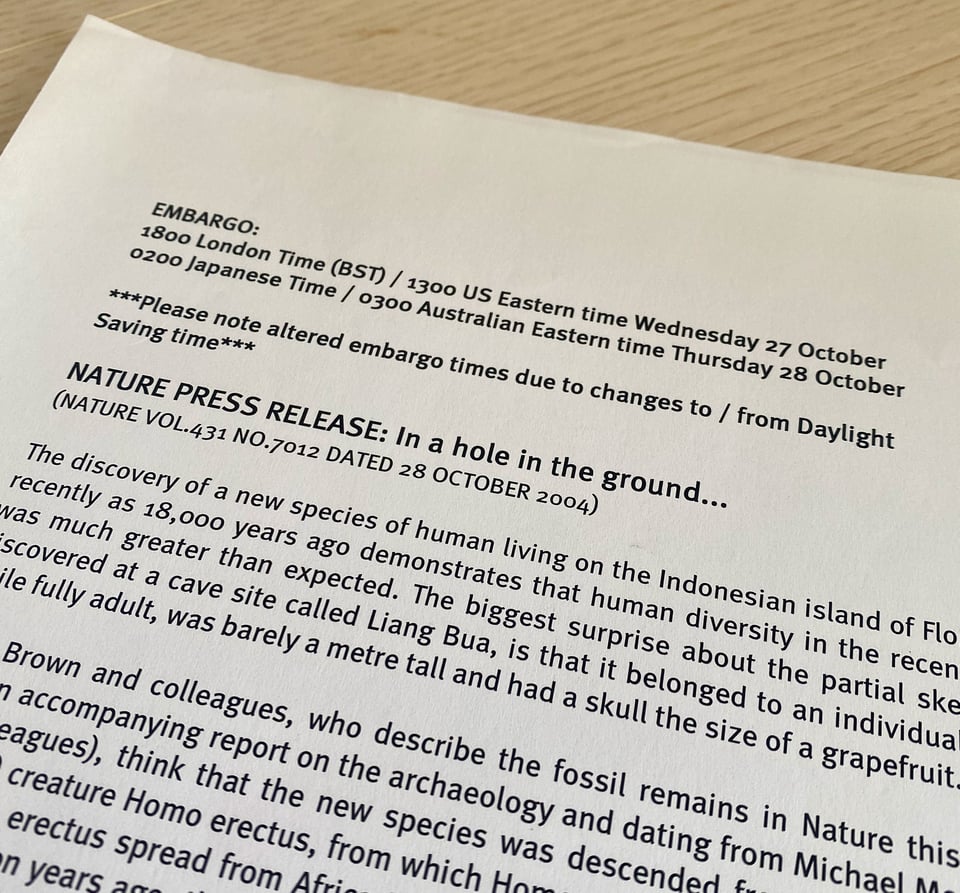 A printed Nature press release lying on a wooden table. The header gives embargo times for Wednesday 27 October, and the title reads “NATURE PRESS RELEASE: In a hole in the ground…”, dated 28 October 2004. The visible text describes the discovery of a new species of human that lived on the Indonesian island of Flores as recently as 18,000 years ago, found at a cave site called Liang Bua.