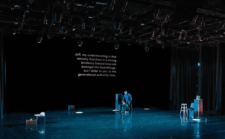 a shot of a large theater with shiny floors, pale blue light, a few people standing or crouching toward the back wall, a few apple boxes stacked or spread about, and a chunk of text projected on the back wall.