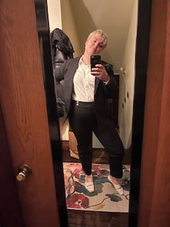 Nola (white bleached blonde short haired femme) in a black and white suit, looking mildly pleased with her reflection in the mirror
