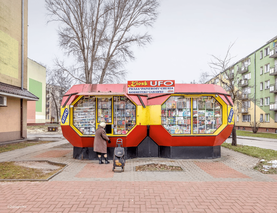 A picture of a yellow and red street stall, with the words "UFO" on its sign. It appears to be selling magazines or candies. A woman in a brown coat is at one of the two octagonal windows