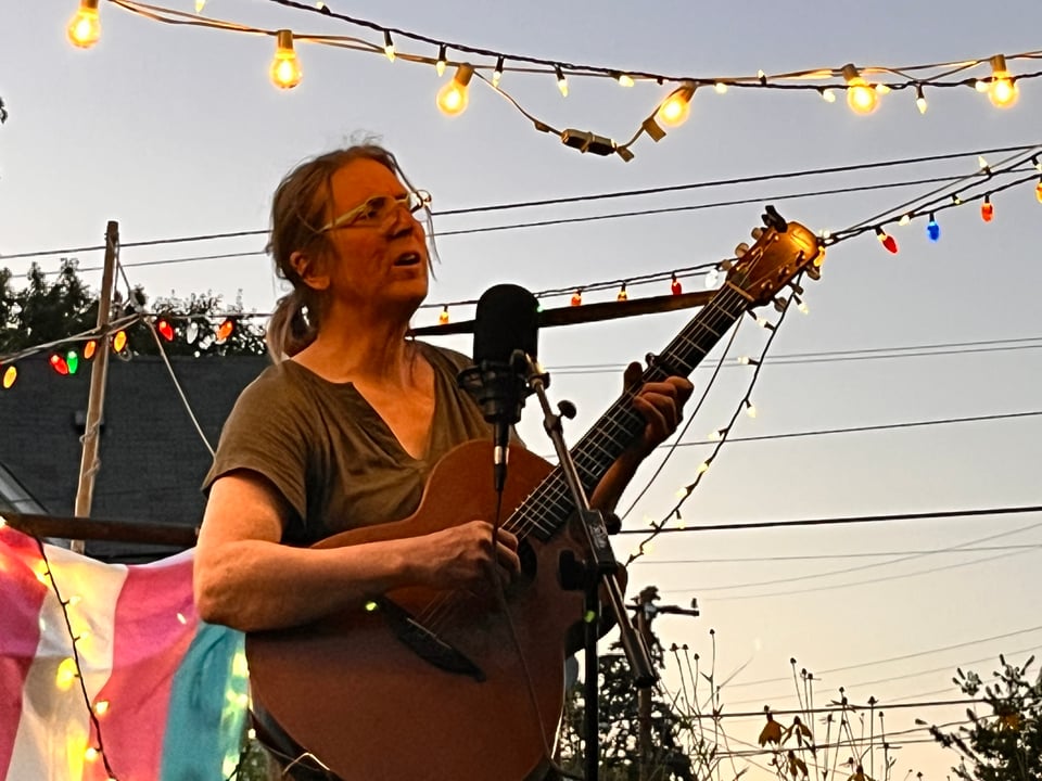 me, on a stage strumming and singing, with a hundred little glowing lights around me, the trans pride flag hung behind me, and a moon faintly rising in the background.