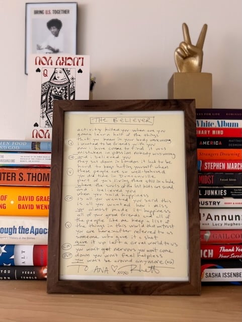 Framed hand-written lyrics displayed on a shelf among books (Hunter S. Thompson, Joan Didion, David Graeber, Stephen Marche, Isabel Wilkerson’s Caste, etc.), along with a peace sign sculpture and some personal cards/prints (including a Queen of Hearts with "Lucky You" printed on it).   The Believer Activity killed you When are you gonna find half of things that you knew in your body were wrong I wanted to be friends with you Now I have come to find it was mistaken In passion nobody was wrong  And I believed you  They sat down in London Had to be hard to be hard to keep hating yourself when these people are so well behaved You did time in Duncanville Part of you is living there still in a hole where the souls of the lost geared to save  And I believed you  (CHORUS) When you said happiness Is all you wanted You said this Is all you wanted How I miss We almost made it Happiness  All of your good friends All of the people like me keep a list of the things in this world that we trust You are here in after referred to as someone who gave it a shot, gave it off, left the cruel world to us