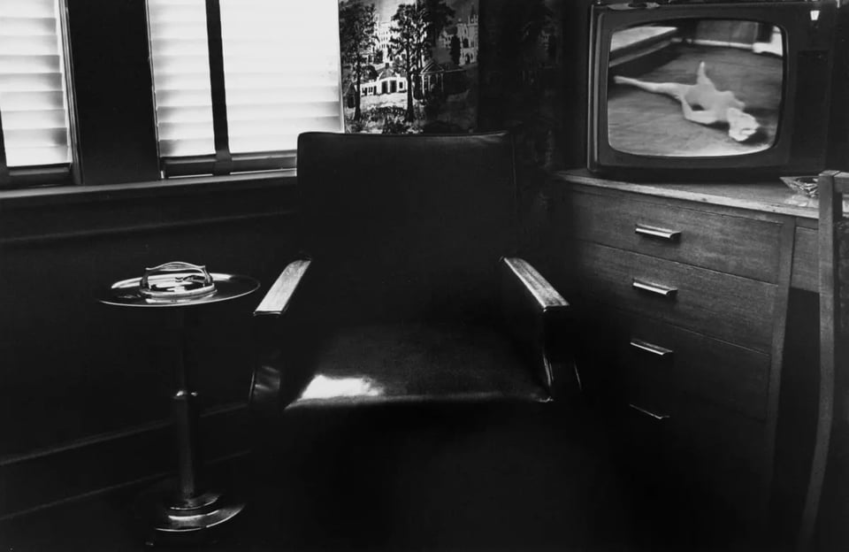 black and white photograph of a hotel room, a chair sits in the corner of the room in front of a window, a stand up ashtray beside it, a television is on in the backgroun with an image of a woman in a leotard lying on the ground