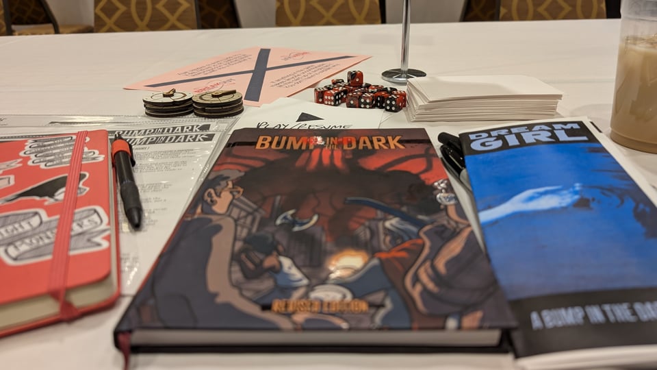A photo of a table with a white tablecloth. On it, in the center of the frame, is a book called Bump in the Dark. A pamphlet next to it reads "Dream Girl: A Bump in the Dark Hunt," and the table also contains a moleskin notebook, some dice, and other TTRPG paraphernalia.