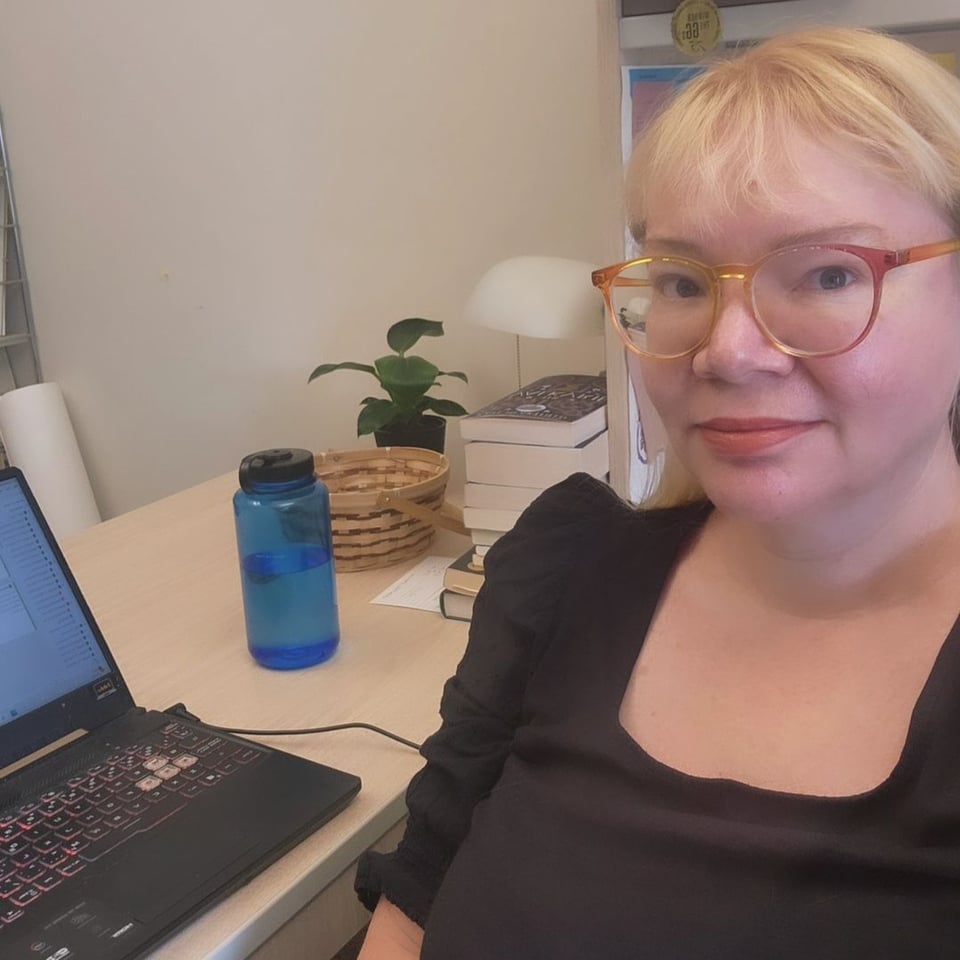 A blonde woman with glasses sits at a desk with an open laptop, a Nalgene water bottle, a basket that contains wrapped candies, a stack of books and a plant.
