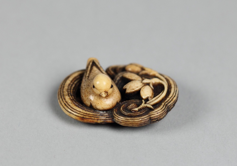 Ivory colored sculpture of a bird sitting on the left side of a ridged, oval nest with a curling flower carved into the right side.