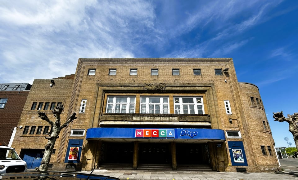 A 1930s honey-coloured brick cinema with Deco detail. The main front is a giant squared arch, with rib detail. A second square arch surrounds large windows set behind a curved balcony. Below that is the Mecca Bingo canopy and three wide double doors leading within.
