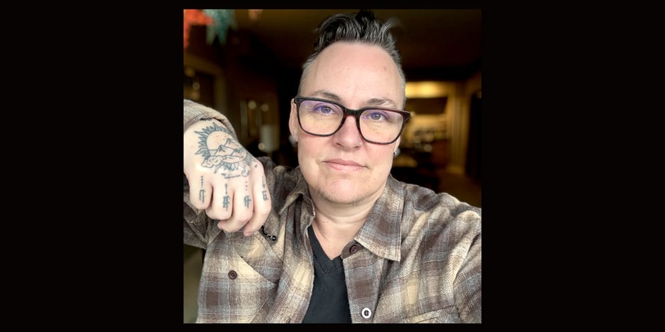 Leoh looking at the camera, wearing glasses and a brown plaid button up shirt showing a tattooed left hand