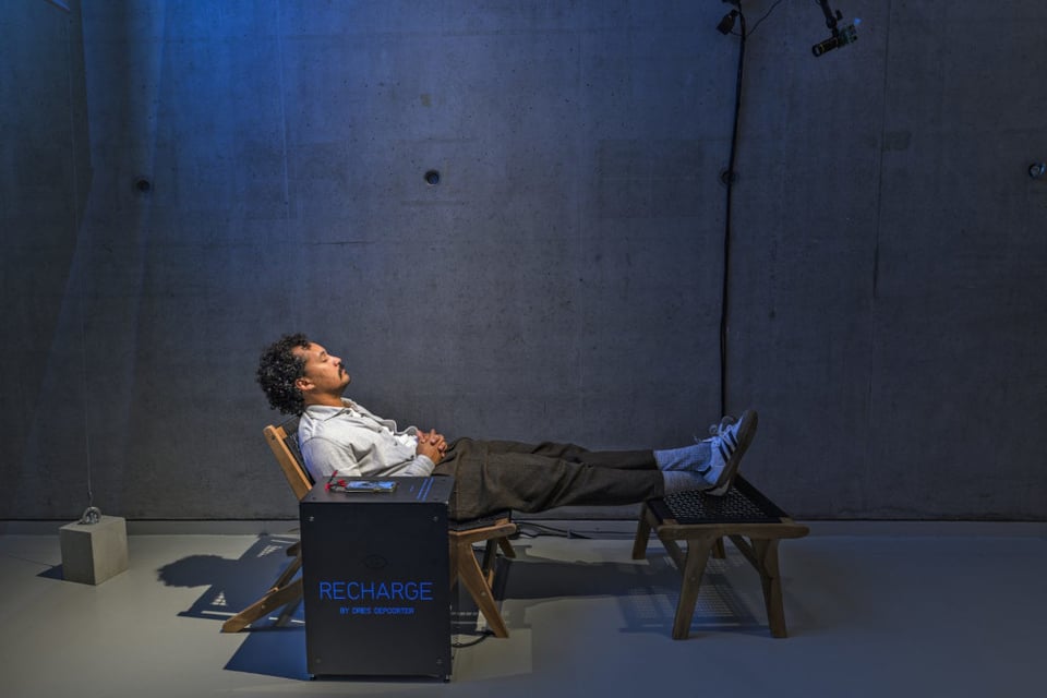 A photo of a man lying down in a recliner chair with his speed up on a footrest. Next to him is a black cube table with the word "recharge" written on it. His phone sits beside him on top of the cube. At the top of the photo on the far right there is a camera sticking down from the roof, pointed at his face. There is a spotlight from the ceiling illuminating the upper half of this body. Otherwise, the room is empty with cement walls and floor, grey.