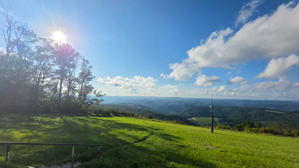 A sunlit hilltop meadow of green grass overlooking rolling forested mountain ridges, with tall trees silhouetted against the bright sun on the left, a thin antenna pole on the right, a metal handrail and steps in the lower left corner, and scattered white clouds across a blue sky.