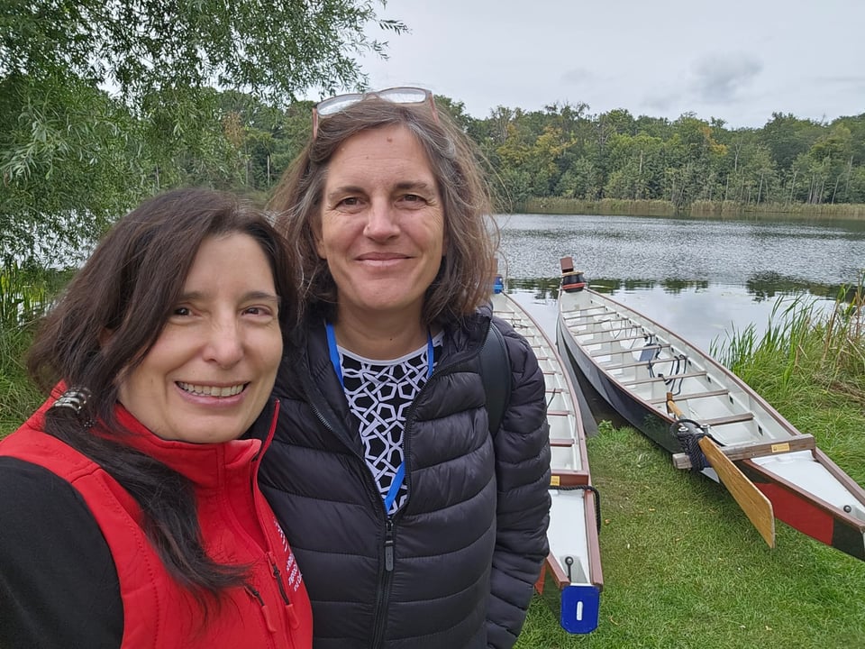 Two women look at the camera as they stand in front of a body of water with two "dragon boats" behind them.