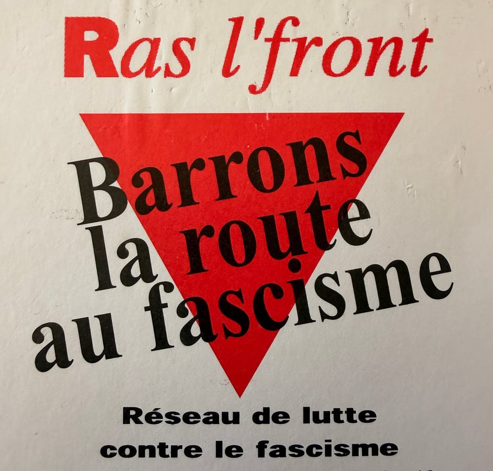 A poster featuring bold text in red and black that reads "Ras l'front Barrons la route au fascisme" within a red triangle, promoting an anti-fascist movement.