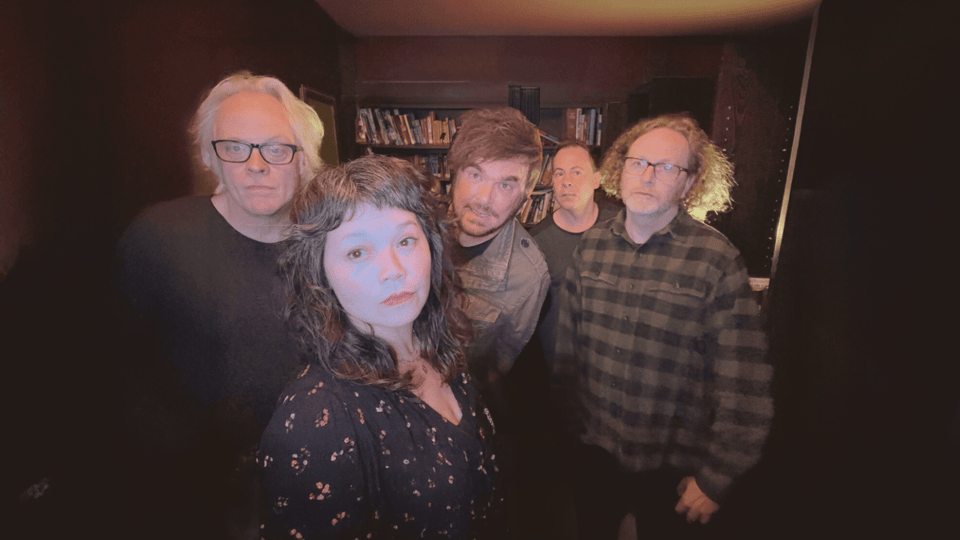 A photo of the band Winterpills, featuring a man on the left with black-rimmed glasses and white-blond hair next to a dark-haried woman and three other men.