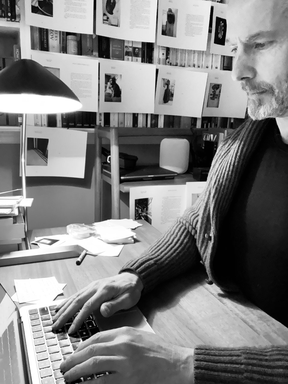 b/w photo of a man in a cardigan and black pullover at a desk, typing on a laptop; pages of a book are taped to the walls behind him