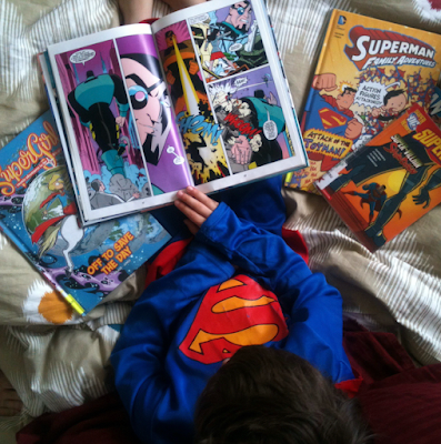 A child in a Superman costume reading comic books while sitting on a bed surrounded by more comics.