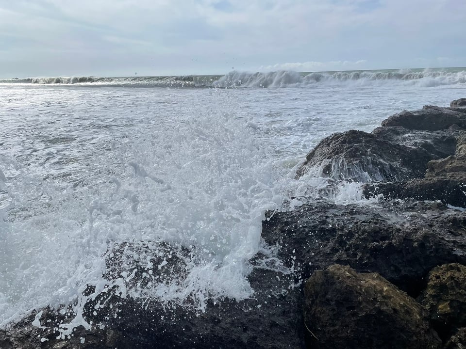 A small wave crashes on the side of the breaker, the splash of water flying directly toward the camera. In the background, the ocean churns and the waves come in