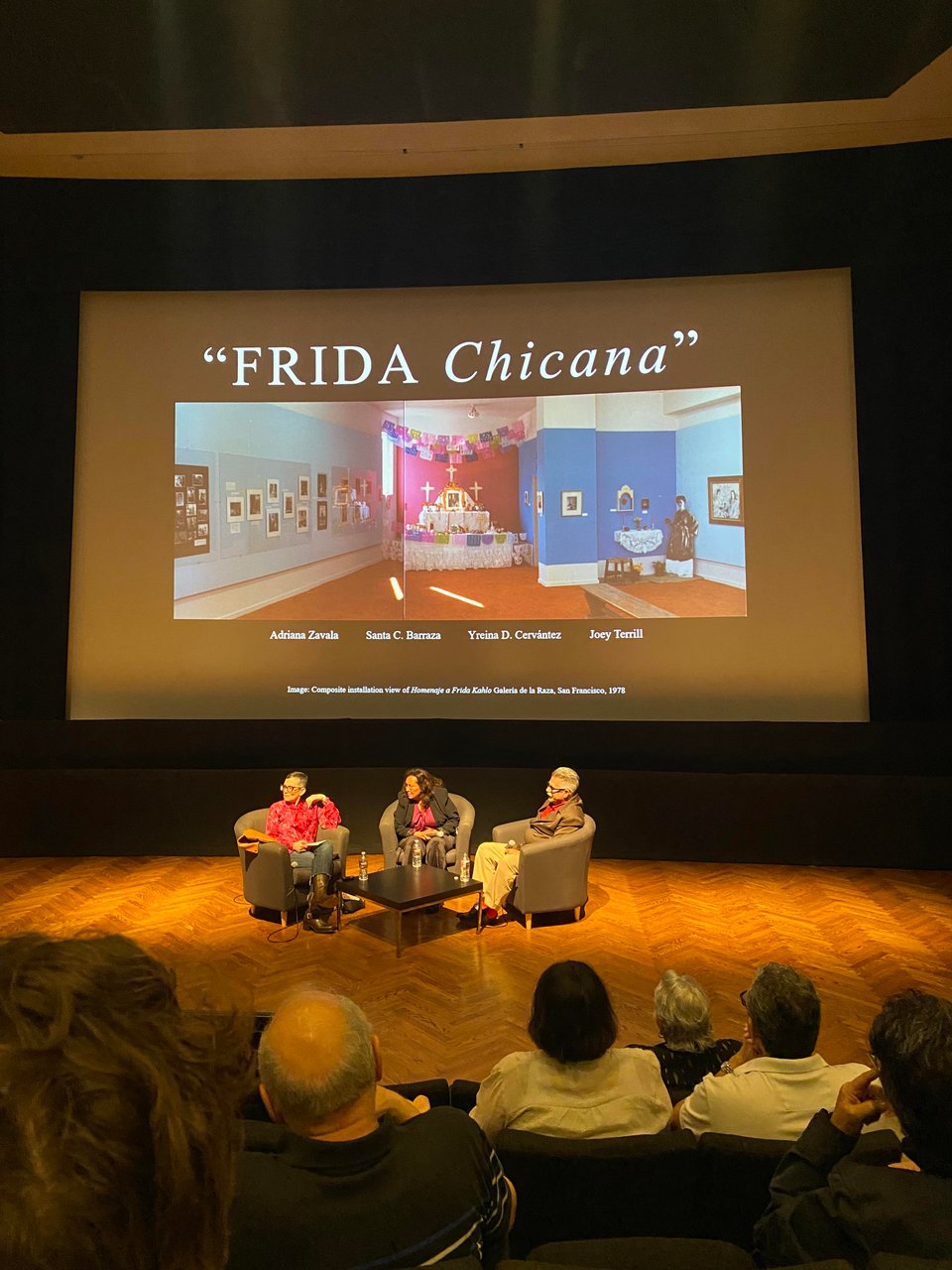 theater view of a panel of artists and scholars in front of a screen that reads Frida Chicana
