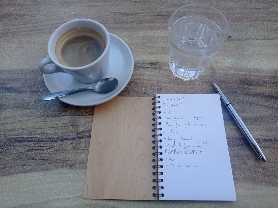 A notebook, a pen, a coffee cup and a glass of water on a coffee table. The notebook has the first lines of Lucie’s scene on its first page. I don’t keep my old notebook so this is a reconstitution made yesterday