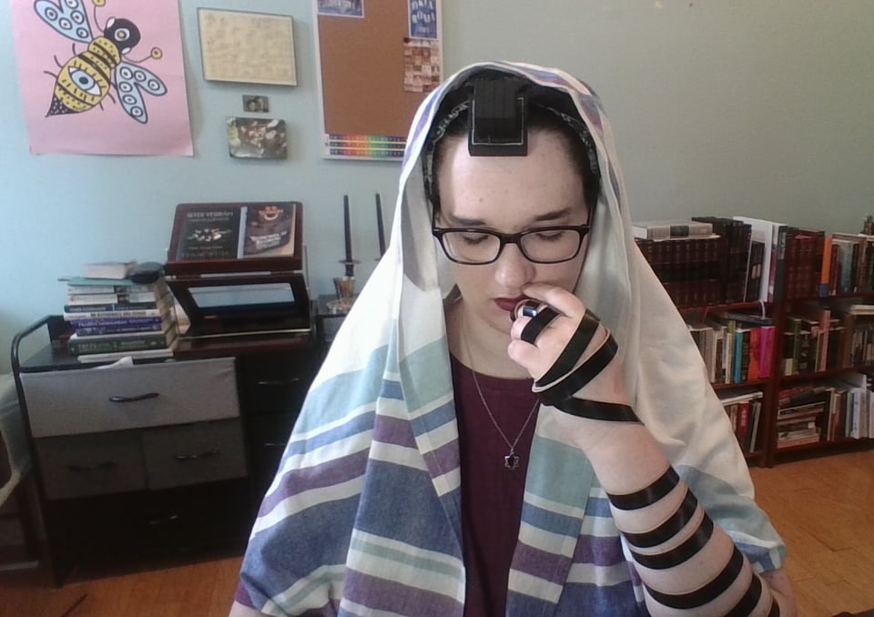 ID: a jewish woman (the author of this post) praying. she is looking down, eyes partly shut. her left hand is raised to her mouth, and has tefillin straps wrapped around it in the shape of a shin. the tefillin straps are visible wrapping up her arm. she is wearing tefillin on her head, covered by a tallis with light pastel bars. she has red lipstick, a dark red shirt, and a magen david necklace. behind her are bookshelves full of jewish books, a nightstand with stacks of books and a shtender with more books on it.