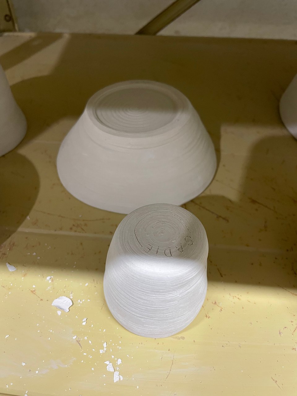 Two unfinished pieces of pottery – a shallow bowl and a cylindrical egg cup – are flipped upside down to dry on a pale yellow shelf. The name Sadie is carved into the bottom.