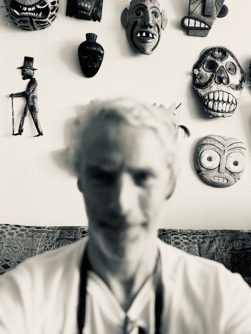 portrait-mode selfie with a blurry white man's face and behind him, in focus, a wall of wooden masks