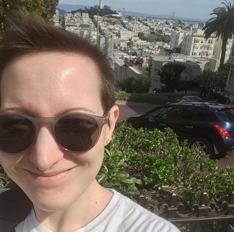 Jess, wearing sunglasses and smiling, stands in front of a view down a sloped street in San Francisco on a sunny day
