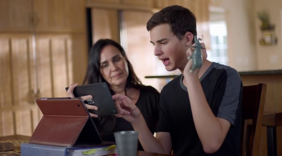 A facilitator sits with a young man who points at an iPad she is holding up before both of them.