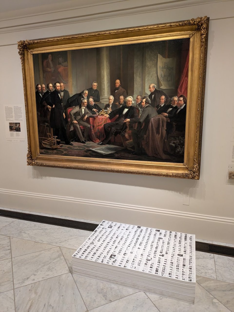 A traditional massive oil painting hangs on the wall, featuring men in formal wear sitting around a table. Below it on the floor is a stack of paper about nine inches tall and 3 by 4 feet in size, featuring tiny photos of people closely packed together.