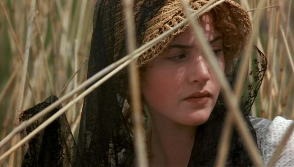 Kate Winslet (a red-haired white woman) as Marianne Dashwood in Ang Lee's Sense and Sensibility. She sits amongst reeds, a black lace scarf of mourning draped over her straw hat, caught on a reed. She looks like an oil painting of an angel.