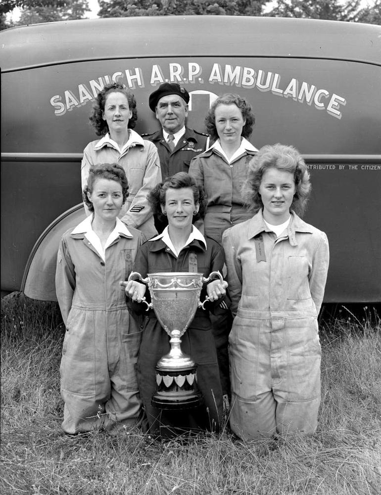 black and white photo of 5 women and one man in front of an ambulance that reads 'Saanich ARP Ambulance." The woman in the front is holding a trophy.