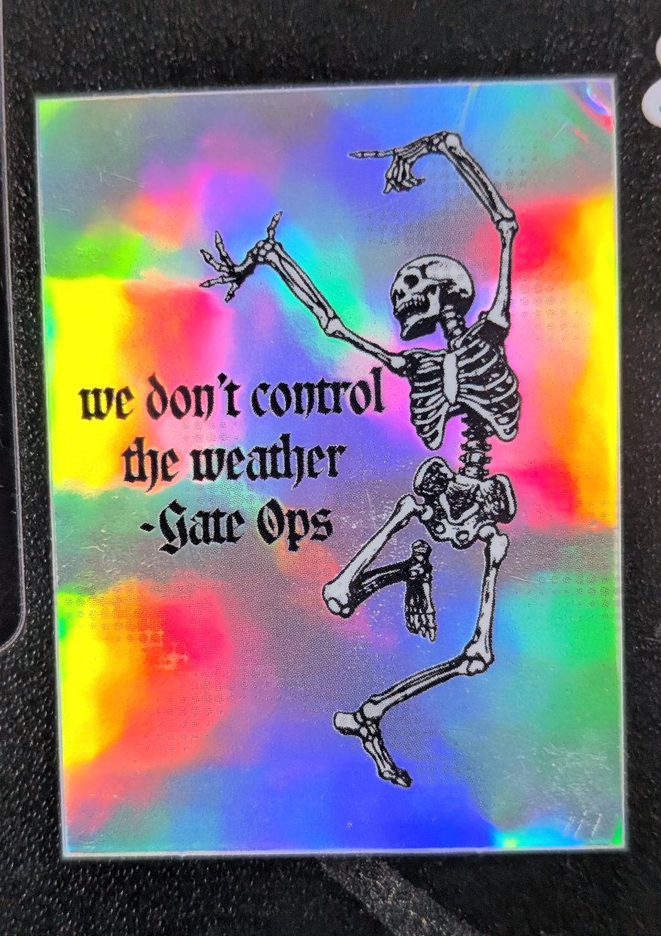 A sticker of a dancing skeleton on a holographic foil background with the text "We don't control the weather -- Gate Ops"