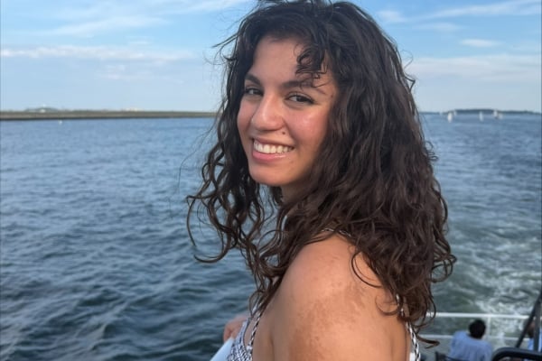 A young woman with curly brown hair smiles brightly while looking over her shoulder from a boat. The background features calm blue water and distant land under a partly cloudy sky.