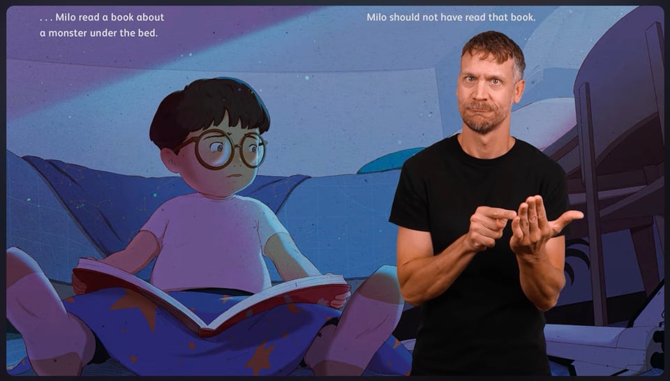 A white male interpreter in black shirt stands in front of an image from the book Monster Hands. It shows a young boy with a book in his lap, glancing nervously around a dark room. The text on the page says, "Milo read a book about a monster under the bed. Milo should not have read that book."