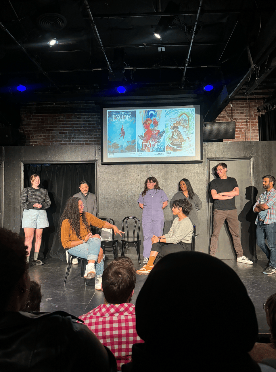 Aabria Iyengar, Kendahl Landreth, Chris Grace, Jordan Myrick, Corin Wells, Grant O'Brien, Sandeep Parikh, and Rekha Shankar on stage in front of a projector with comic book images