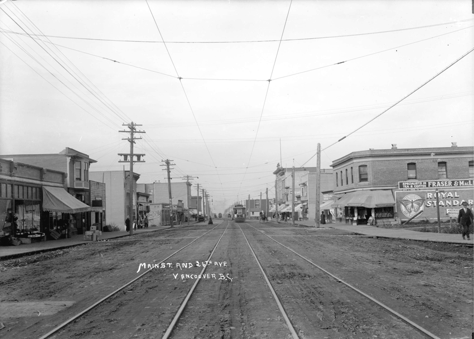 View of a wide dirt road with streetcar tracks. Stores are visible on both sides.