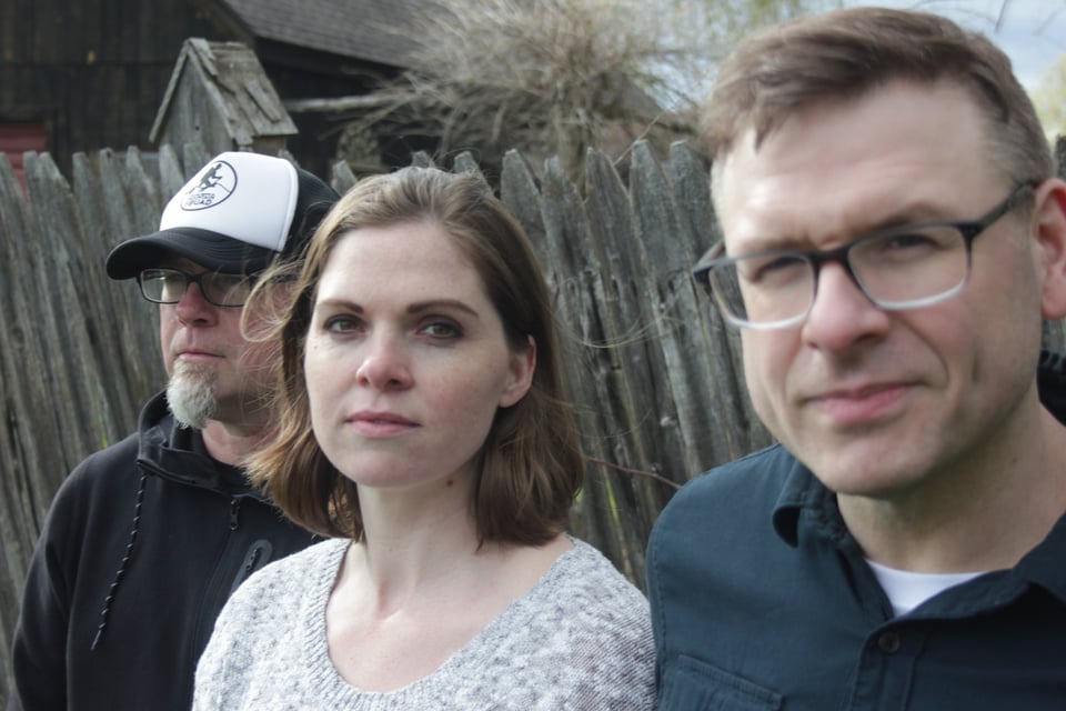 A man in a black and white baseball cap, a woman with shoulder-length brown hairin a gray sweater and a man with short, sandy blond hair and glasses stand together in front of a wooden fence.