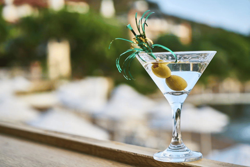 A clear martini glass filled with a pale cocktail sits on a wooden ledge outdoors, garnished with two green olives on a pick decorated with shiny green tinsel. The background is softly blurred, showing sunlit trees and white umbrellas, suggesting a relaxed resort or seaside setting. The image conveys a summery, leisurely mood.