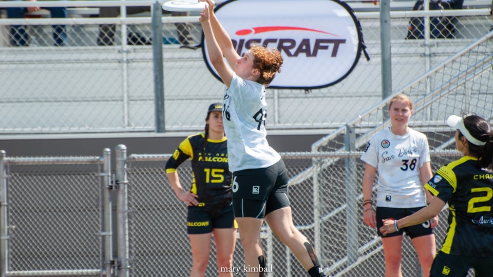 A player in a white jersey catches the disc as their teammate looks on. In the background are bleachers and fences.
