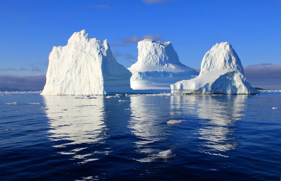 Three icebergs floating, far in the distance, on a dark blue sea. The sky is deep blue behind them