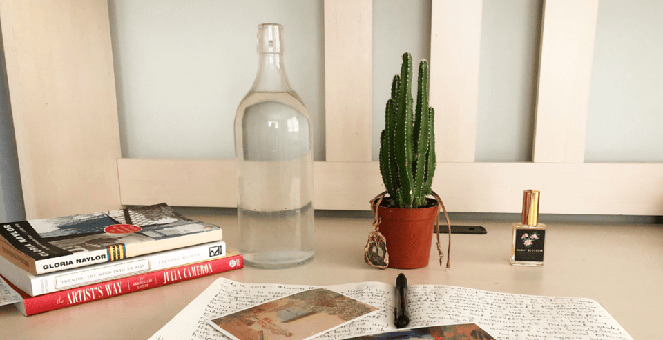 a cream-colored desk showcasing an open notebook with writing on it, a glass bottle of water, a stack of books and a cactus plant.