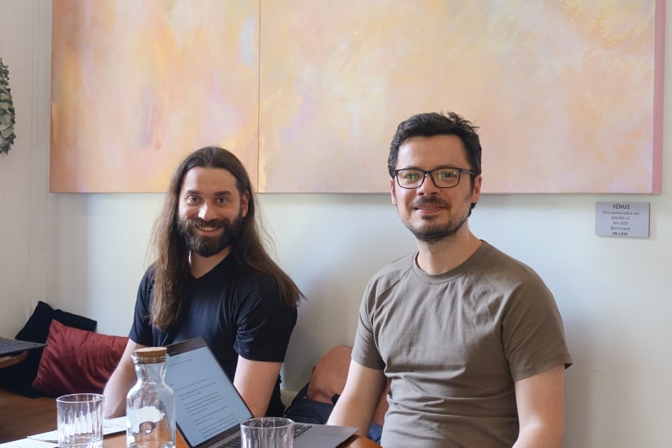 Dois homens sentados à mesa em um café, com um quadro abstrato ao fundo.